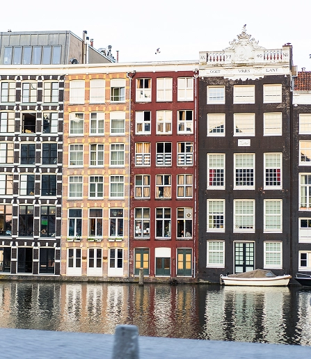 Traditionele smalle grachtenpanden in Amsterdam langs rustige weerspiegelingen aan het water, met klassieke Nederlandse architectuur met hoge gevels, grote ramen, decoratieve daklijnen en aangemeerde boten op de voorgrond, die de historische sfeer van het stadscentrum weergeven